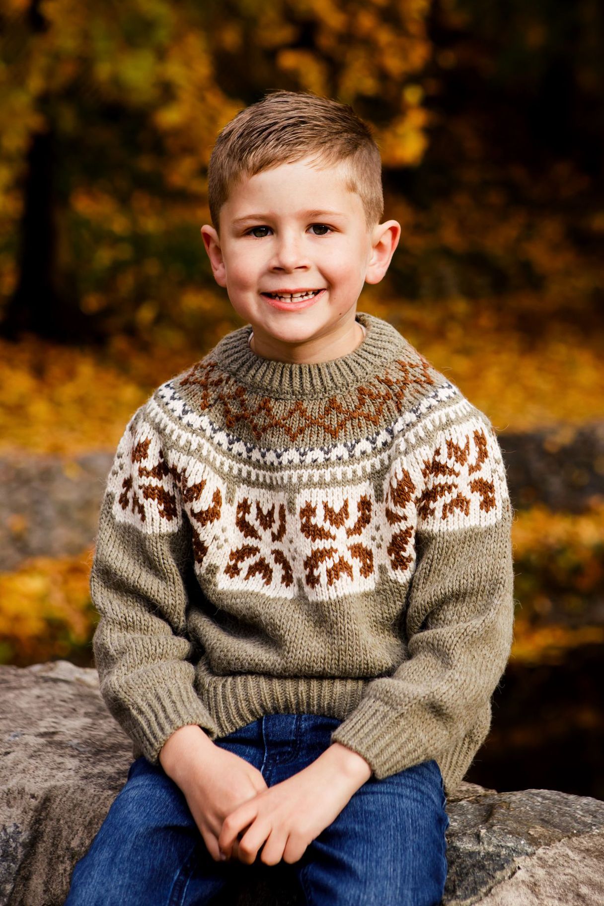 Smiling young boy in a patterned knit sweater sits on a rock in an autumn forest.