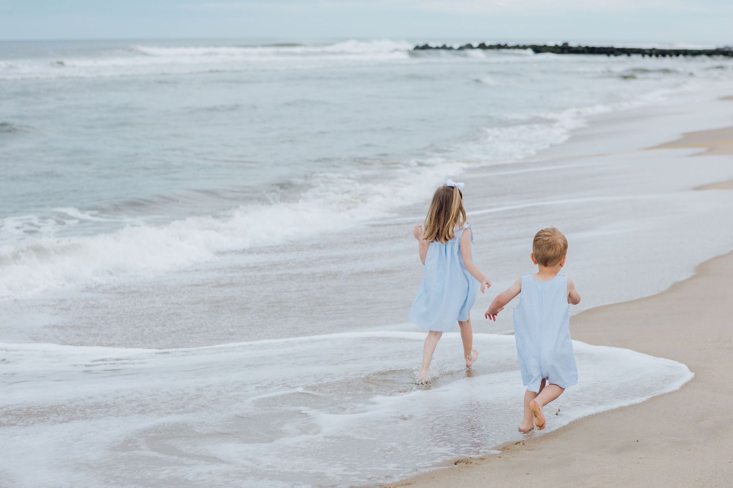 Two little girls in light blue dresses holding hands and walking together along a beautiful ocean beach.