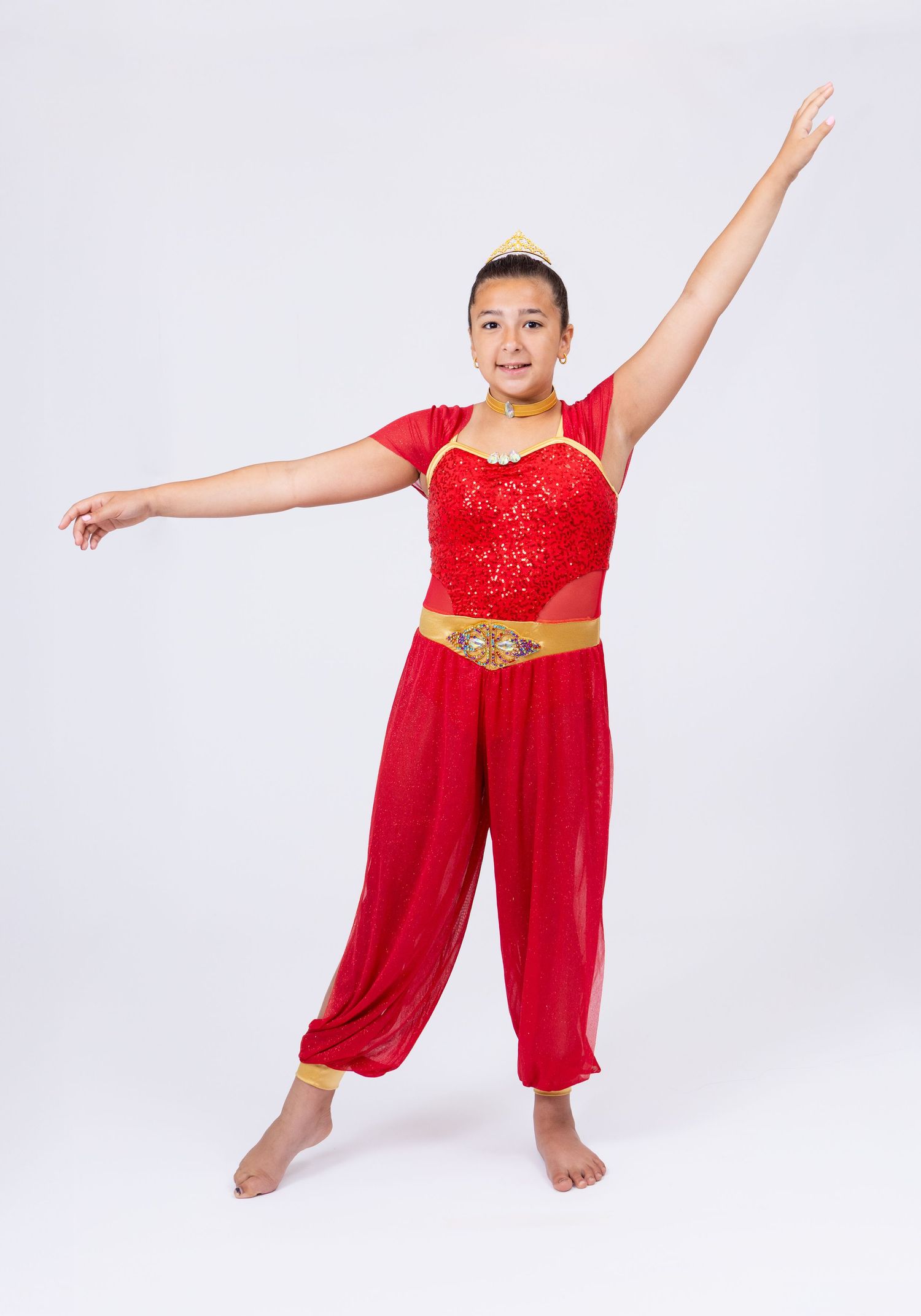 A young dancer performs in a red sequin and gold costume with harem pants in a dance pose.