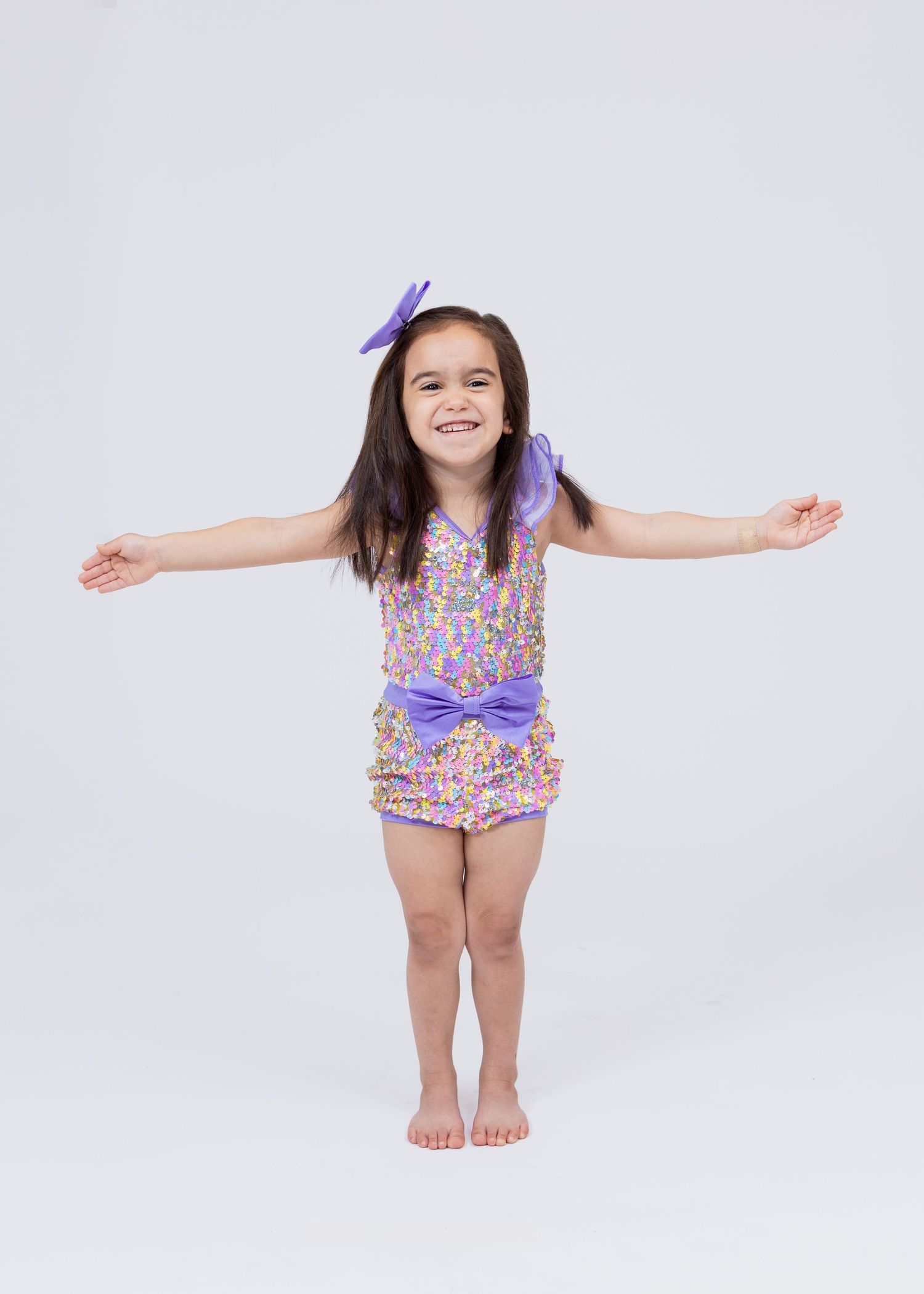 Young girl in purple floral romper with bow hair clip poses with arms outstretched, smiling.