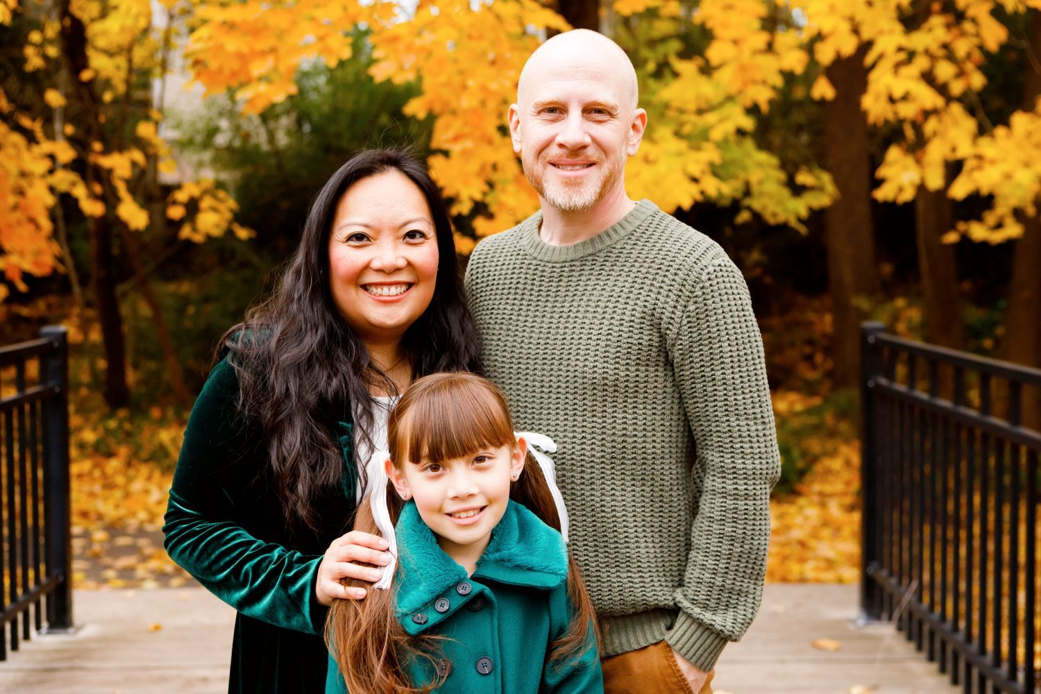 Family of three smiling together on a bridge with vibrant orange autumn trees behind them.