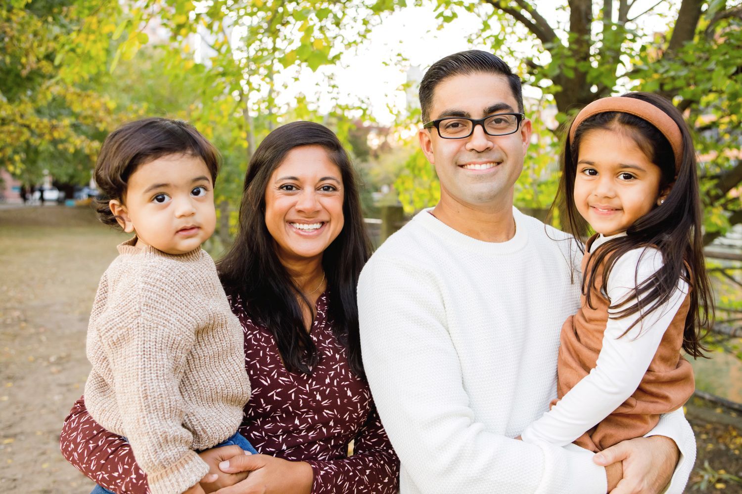 A smiling South Asian family of four posing together outdoors in a colorful autumn park.