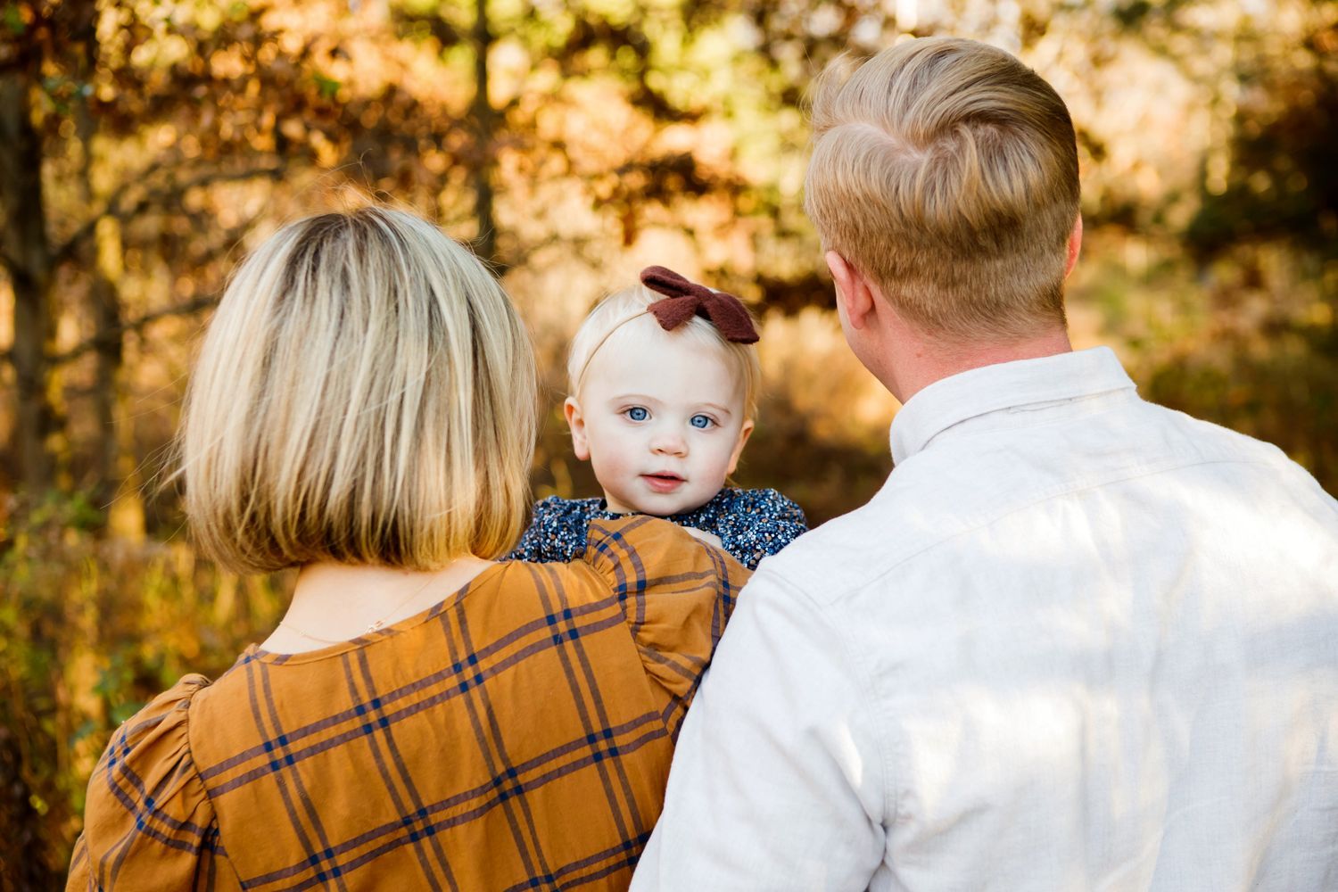 Parents holding their baby girl with a bow headband amid golden autumn forest scenery.