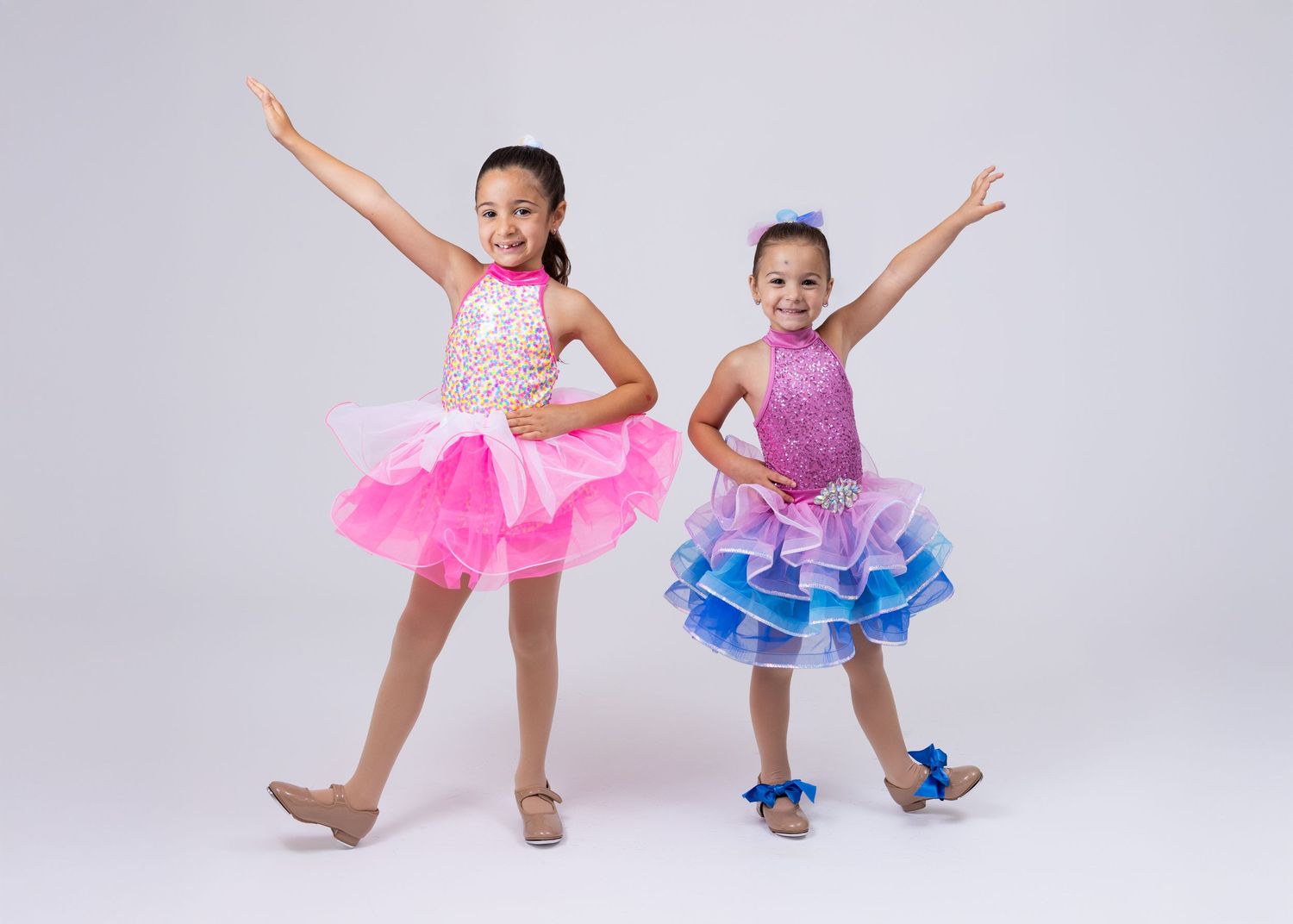 Two young girls in colorful sequined dance costumes striking joyful poses against a white background.