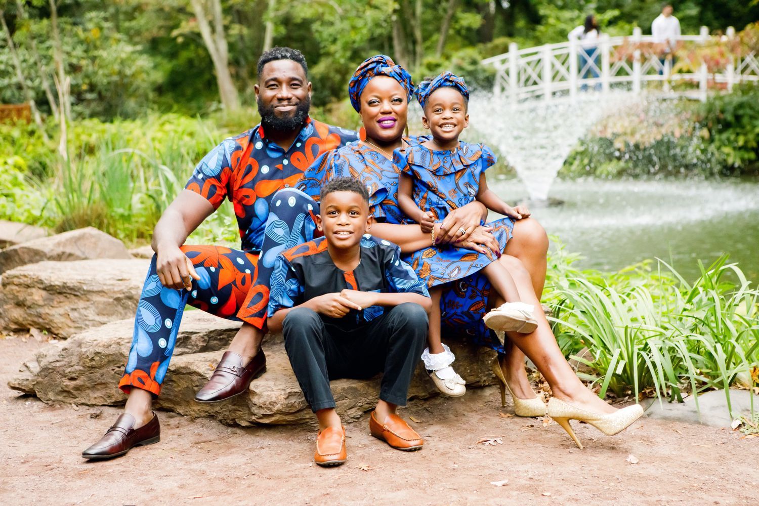 African family of four in matching colorful traditional print outfits poses by a garden bridge.