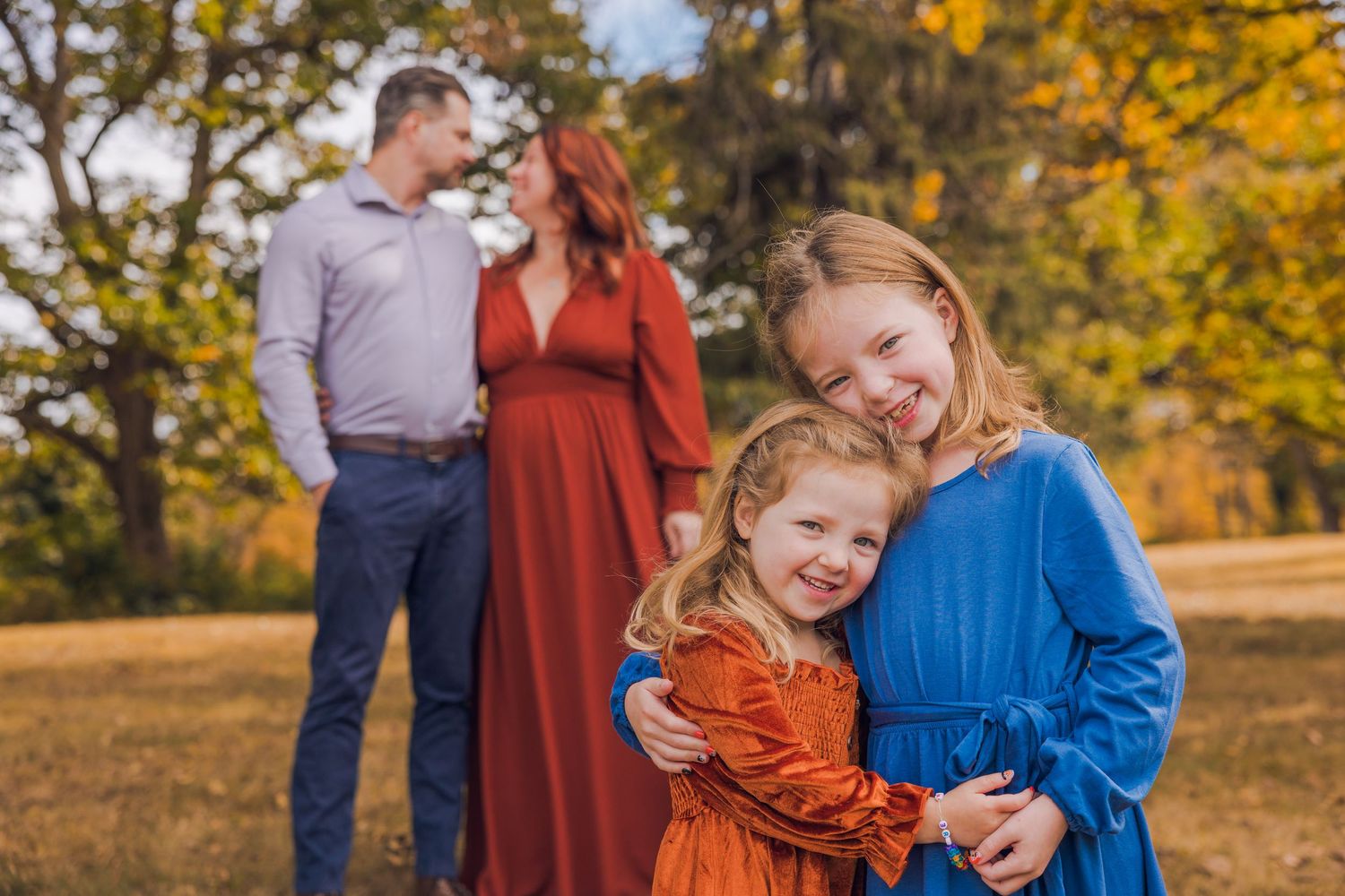 Two young sisters hugging while parents share a romantic moment in a fall foliage setting.