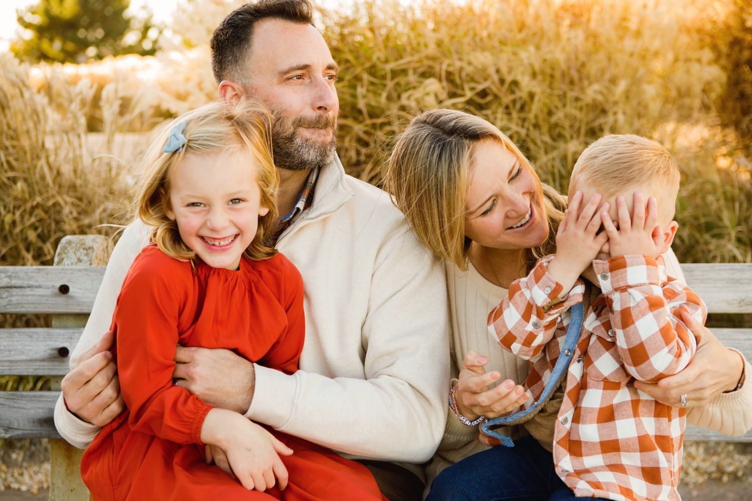 Happy family of four sitting on a bench outdoors during golden hour, parents embracing their two young children.