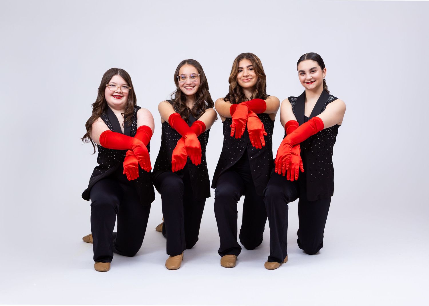 Four dancers in black outfits with red gloves kneel together in a dance studio portrait.