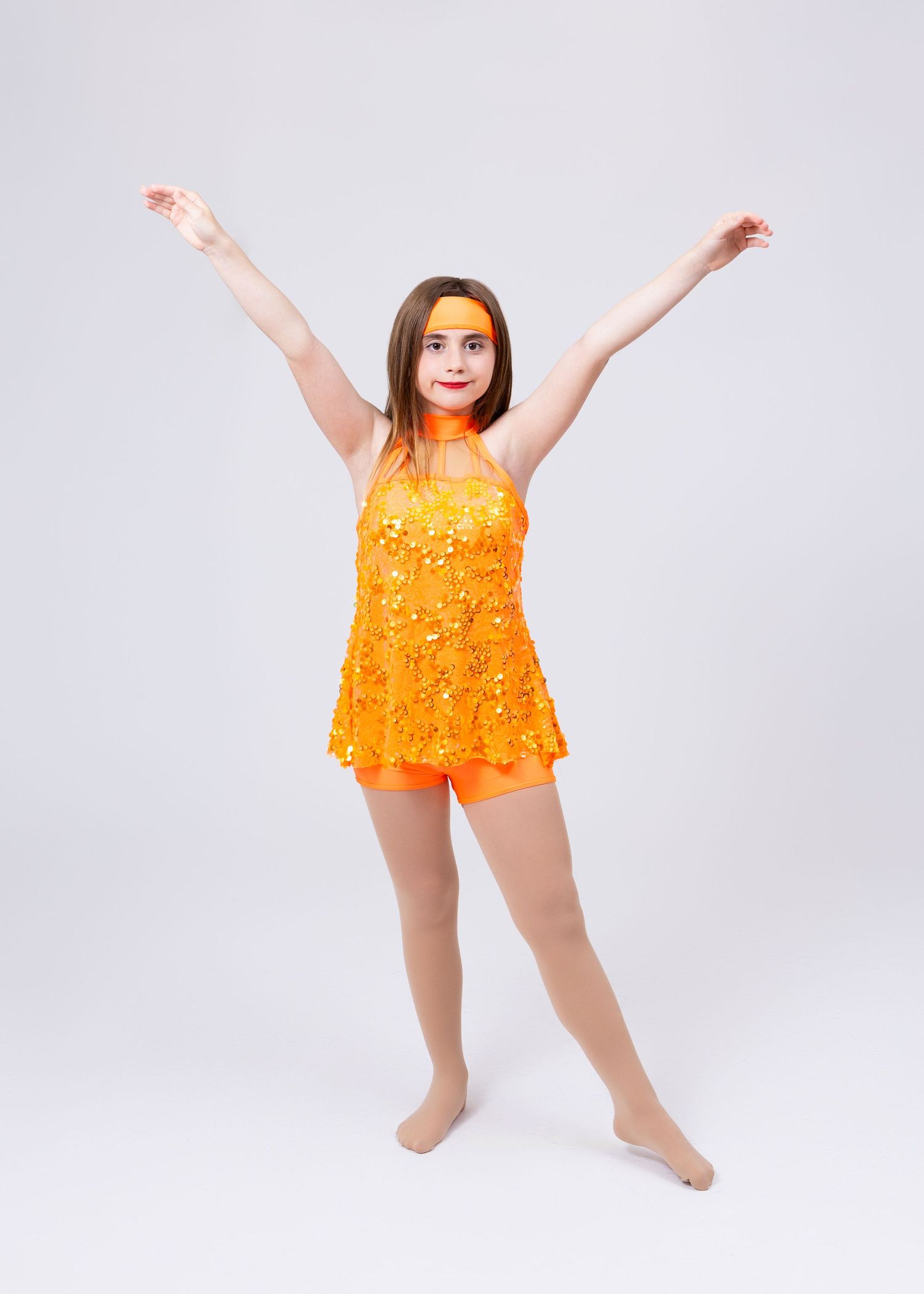 A young girl in an orange sequined dance costume raises her arms joyfully with orange face paint.