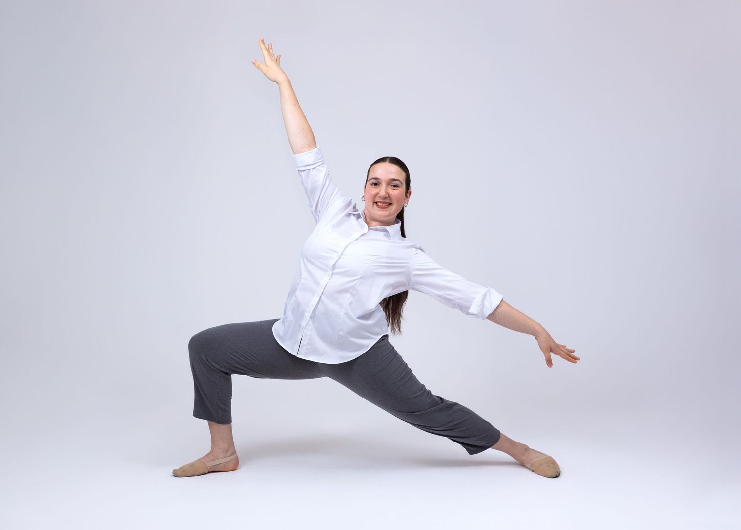 A female dancer in a white shirt and gray pants performs a wide lunge with arm raised in a studio.