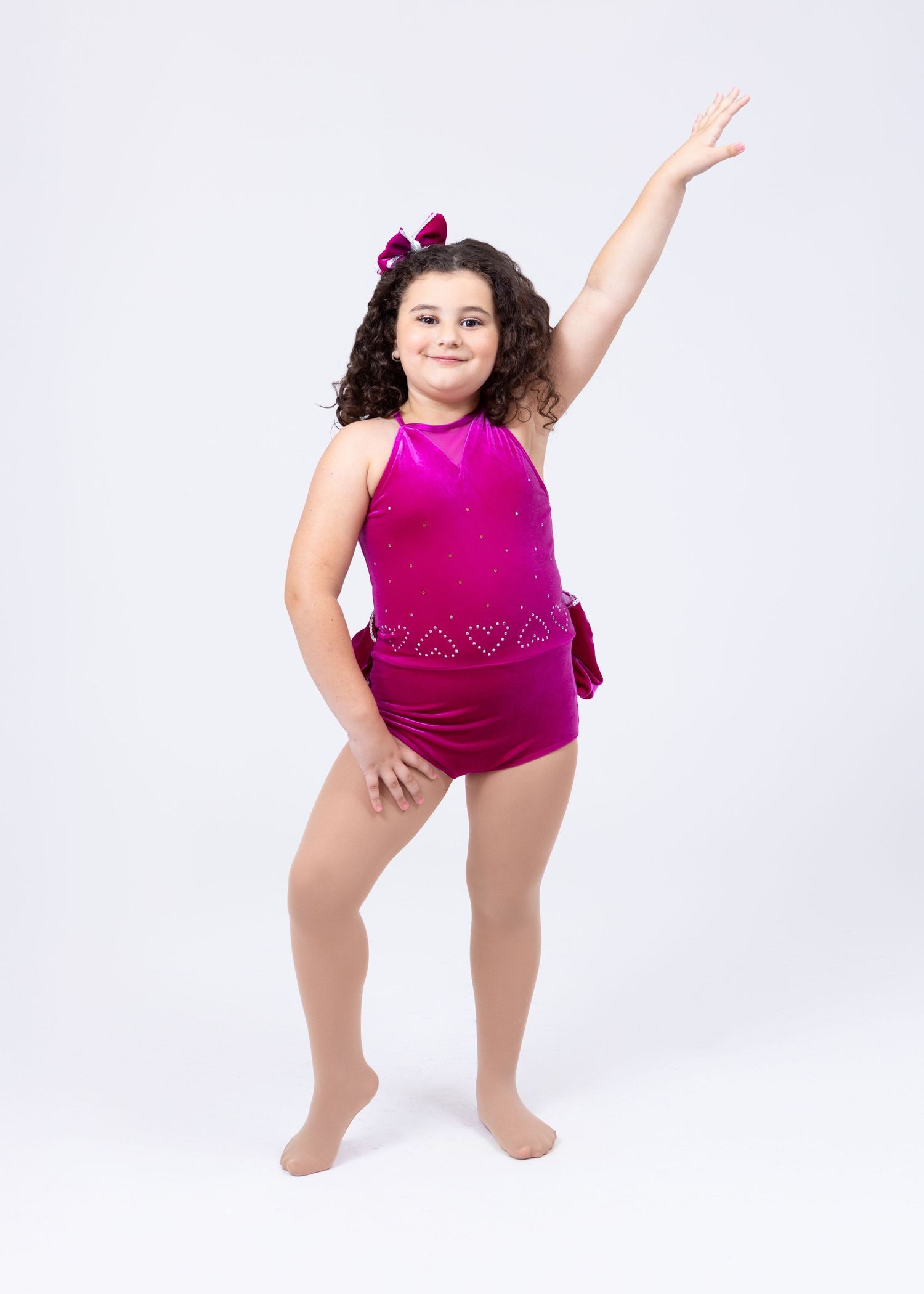 Young girl in magenta sequined dance leotard striking a pose with arm raised on white background.