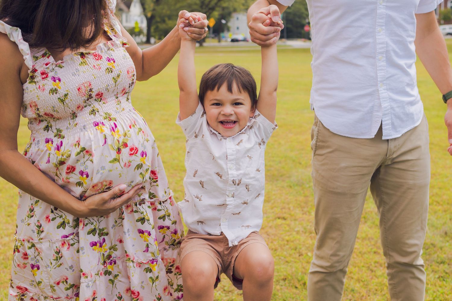 Two parents swing their laughing toddler by the hands on a sunny green lawn outdoors.