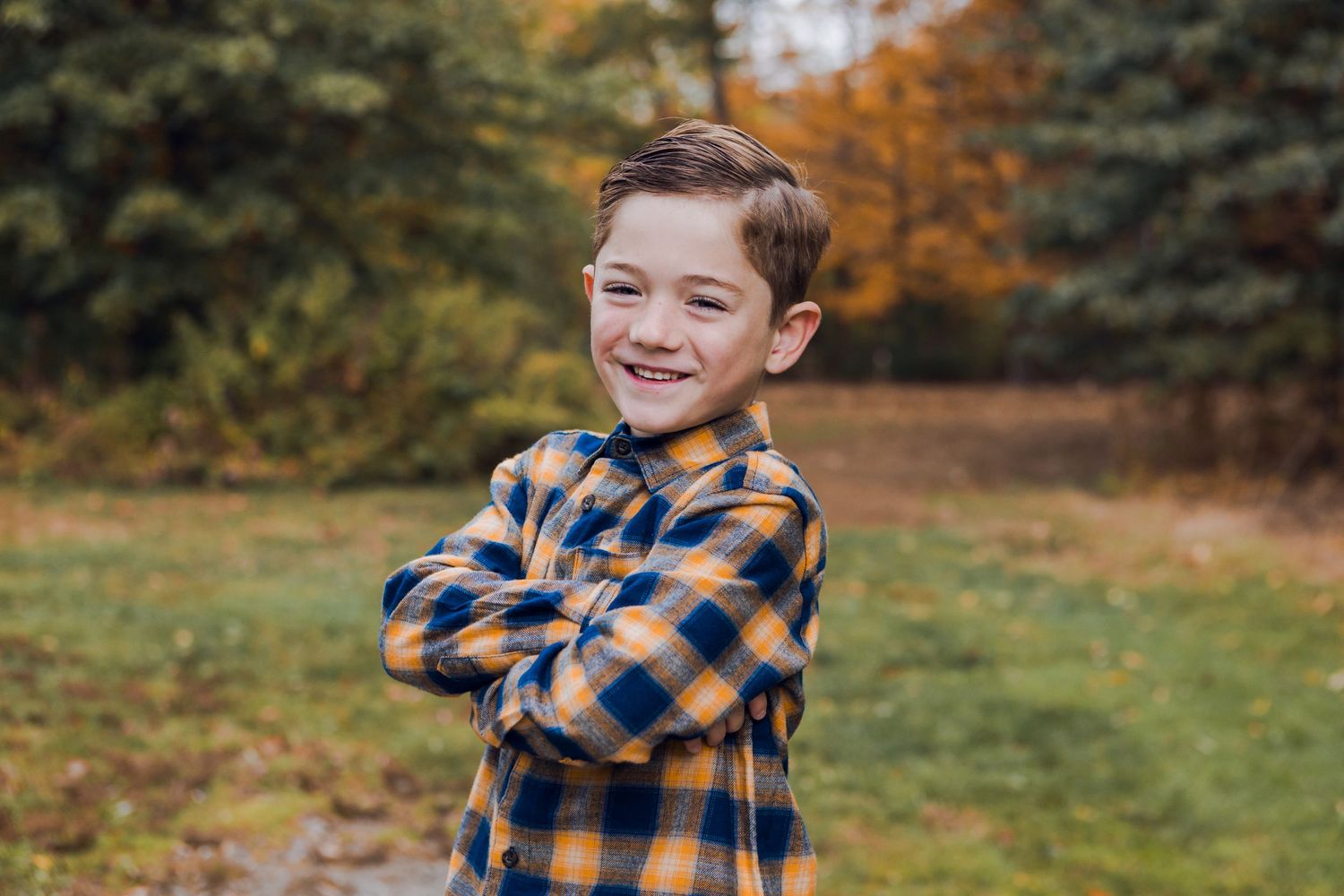 A smiling boy in a blue and yellow plaid shirt stands with arms crossed in an autumn outdoor setting.