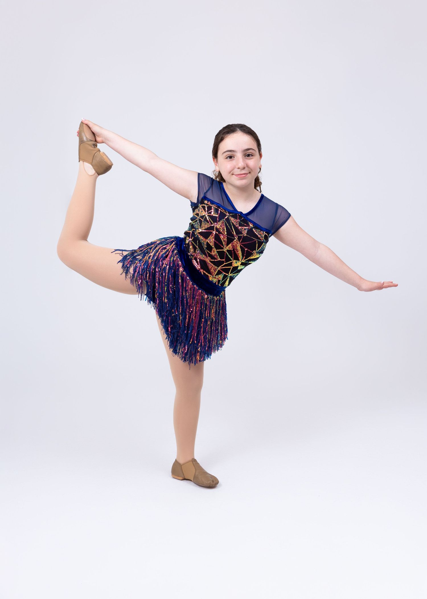 A young girl in a navy sequined fringe dance costume performs a standing leg extension balance.
