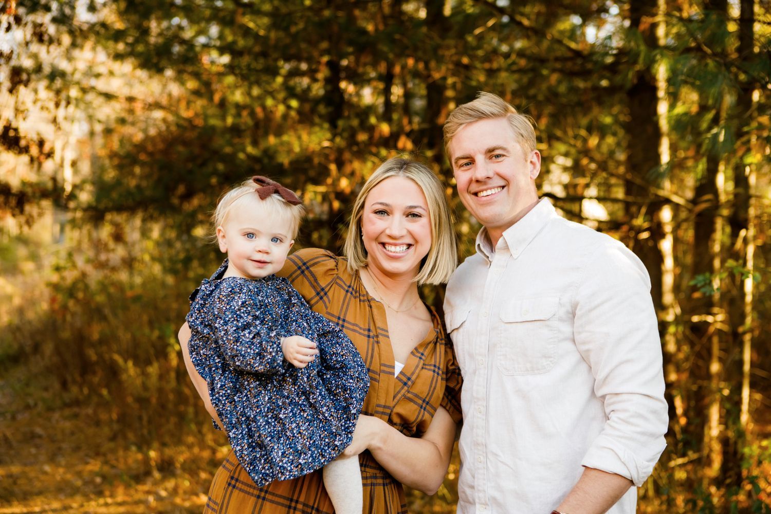 Smiling young family of three posing together on a sunny autumn forest path.