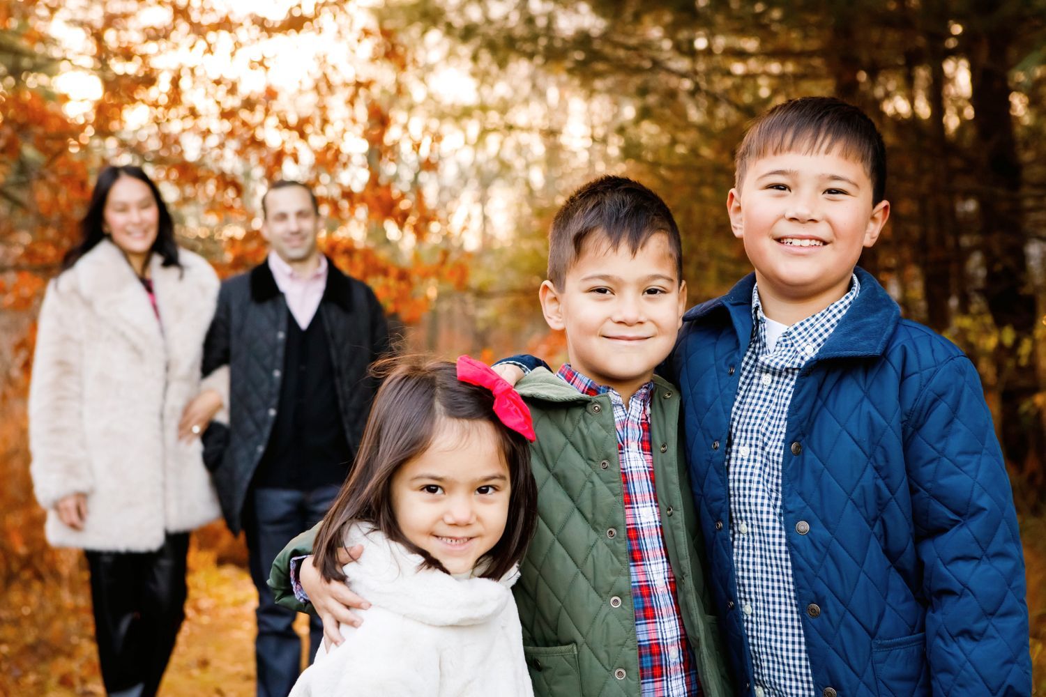 Family of five posing in autumn forest, two parents in background, three children smiling in foreground.
