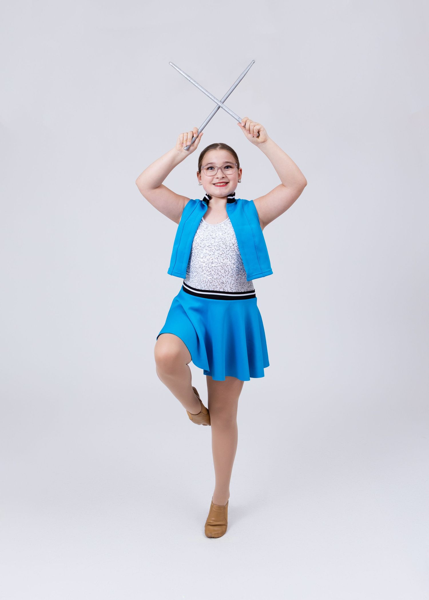 Young girl in blue dance costume balances on one leg while holding two batons crossed overhead.