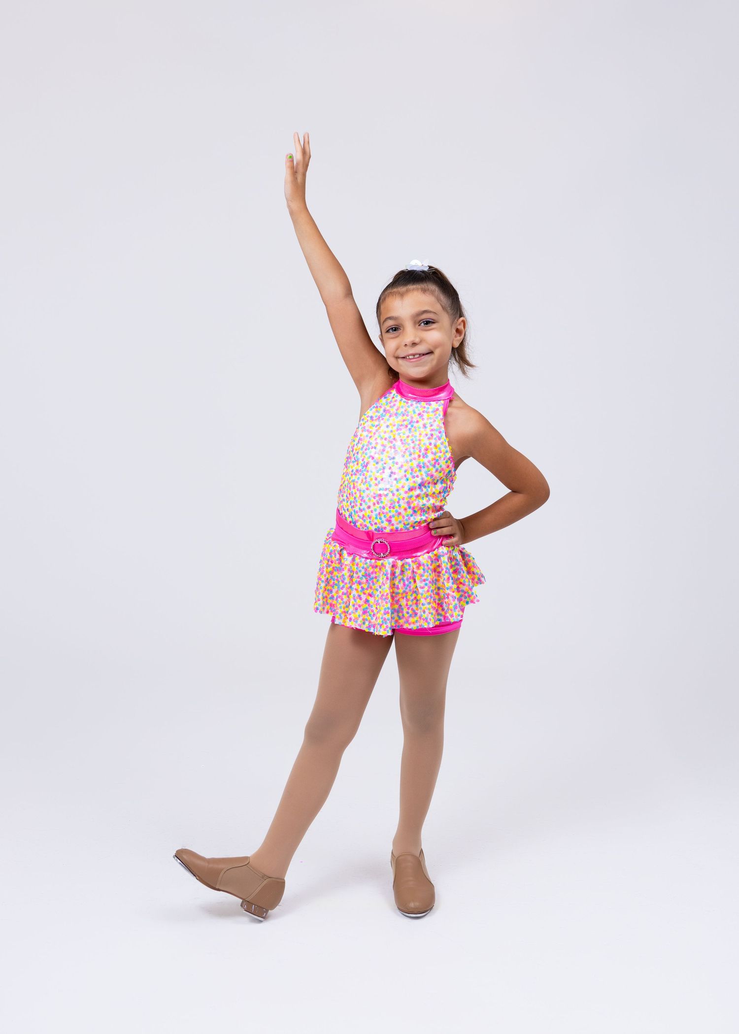 A young girl dancer in a colorful pink leotard and skirt poses with one arm raised in a studio.