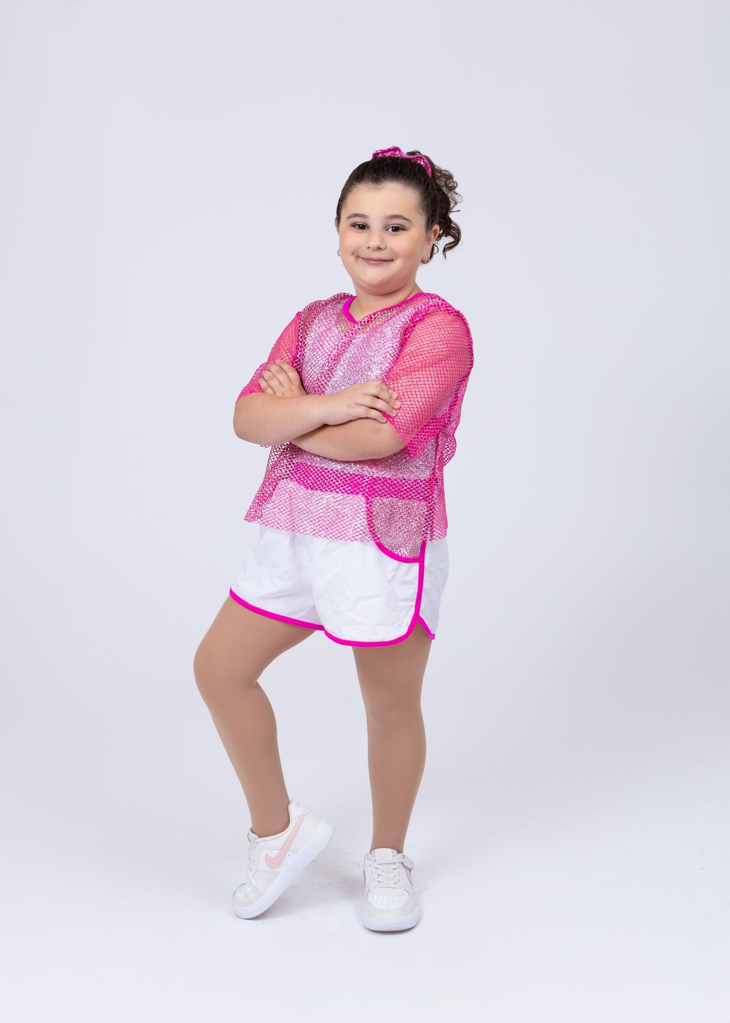 Young girl in pink mesh top and white shorts posing with crossed arms on white background.