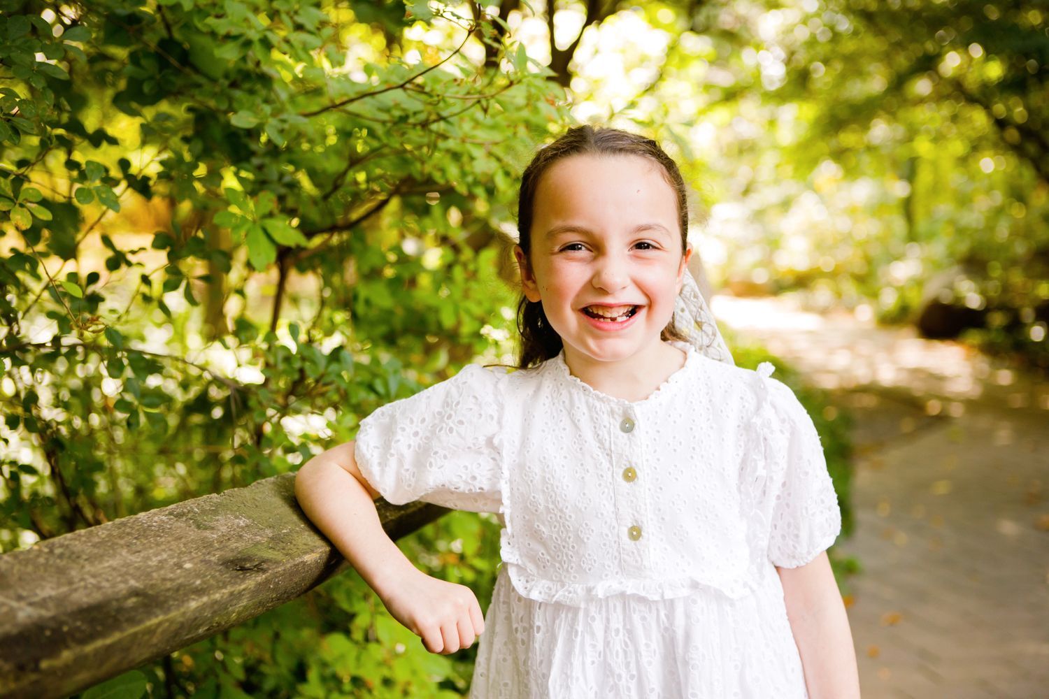 Happy young girl in white eyelet dress laughs while leaning on wooden fence in green park.