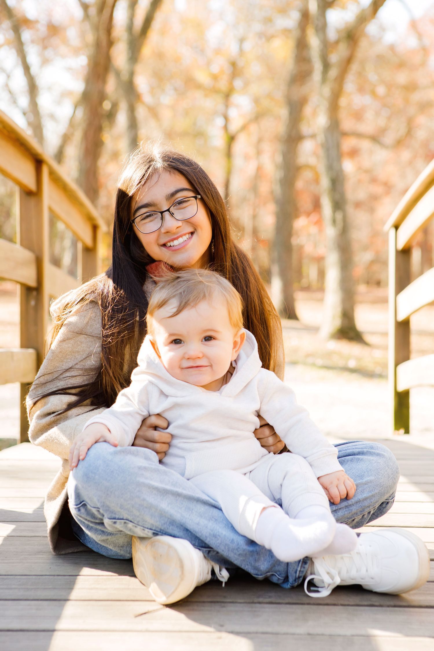 Smiling teenage girl with glasses sitting on a wooden bridge holding a happy baby in an autumn park.