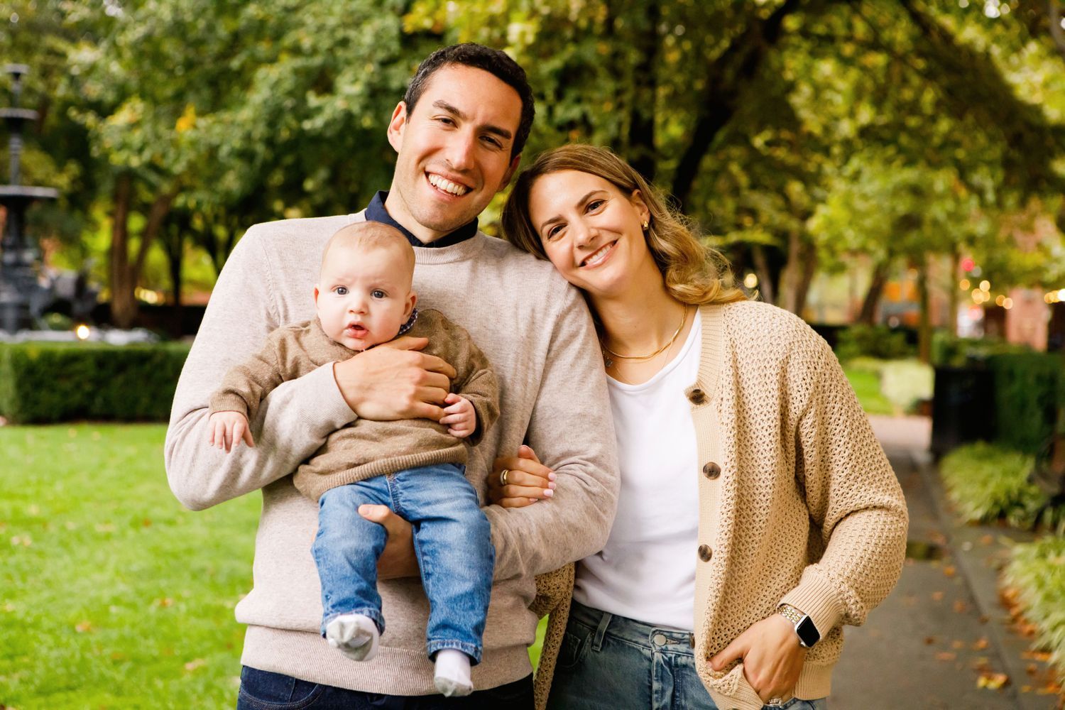 Smiling couple holding their baby outdoors in a lush green park during autumn.