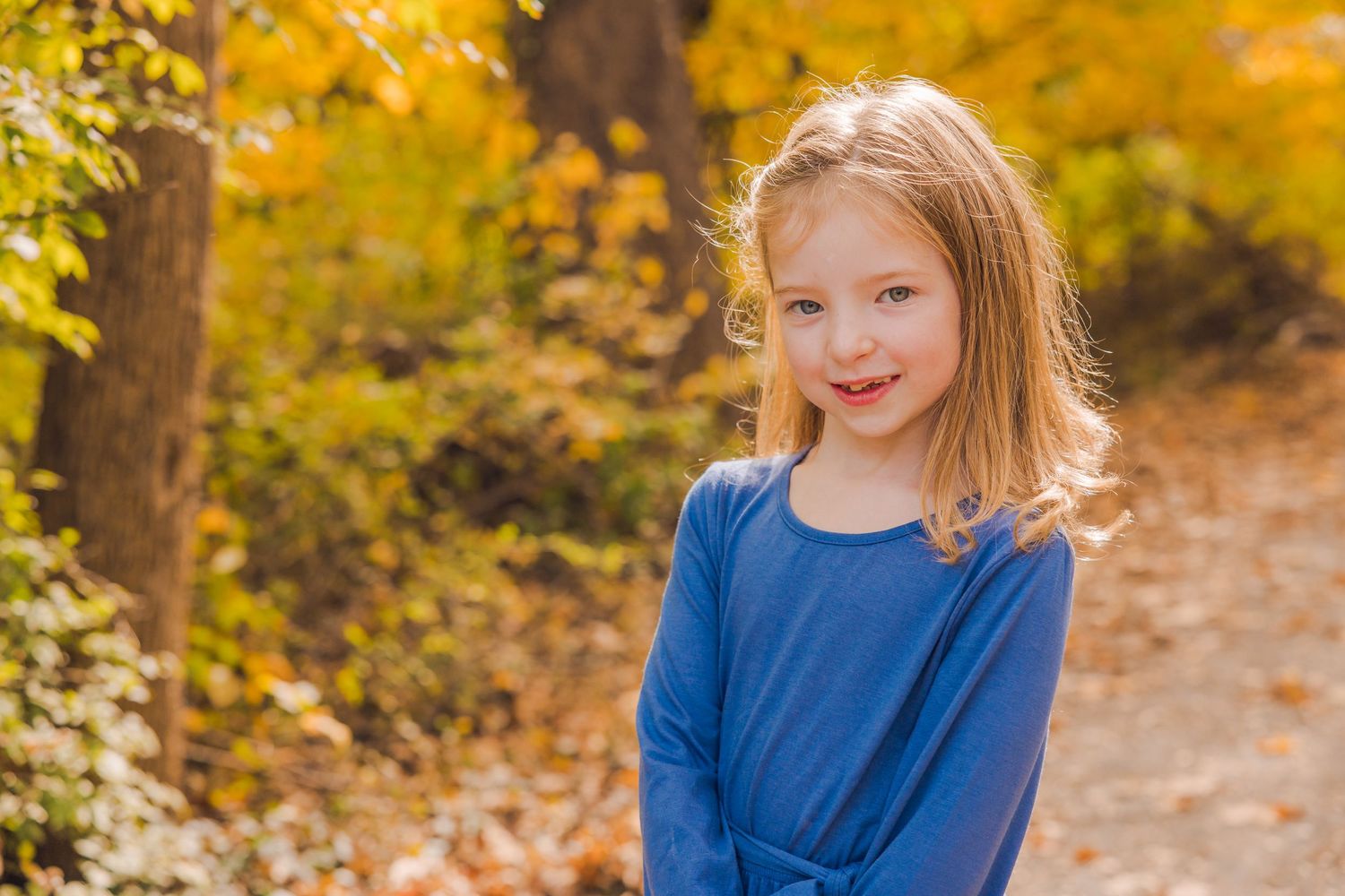 Sweet young girl in blue top smiling in a sunlit autumn forest with golden leaves.