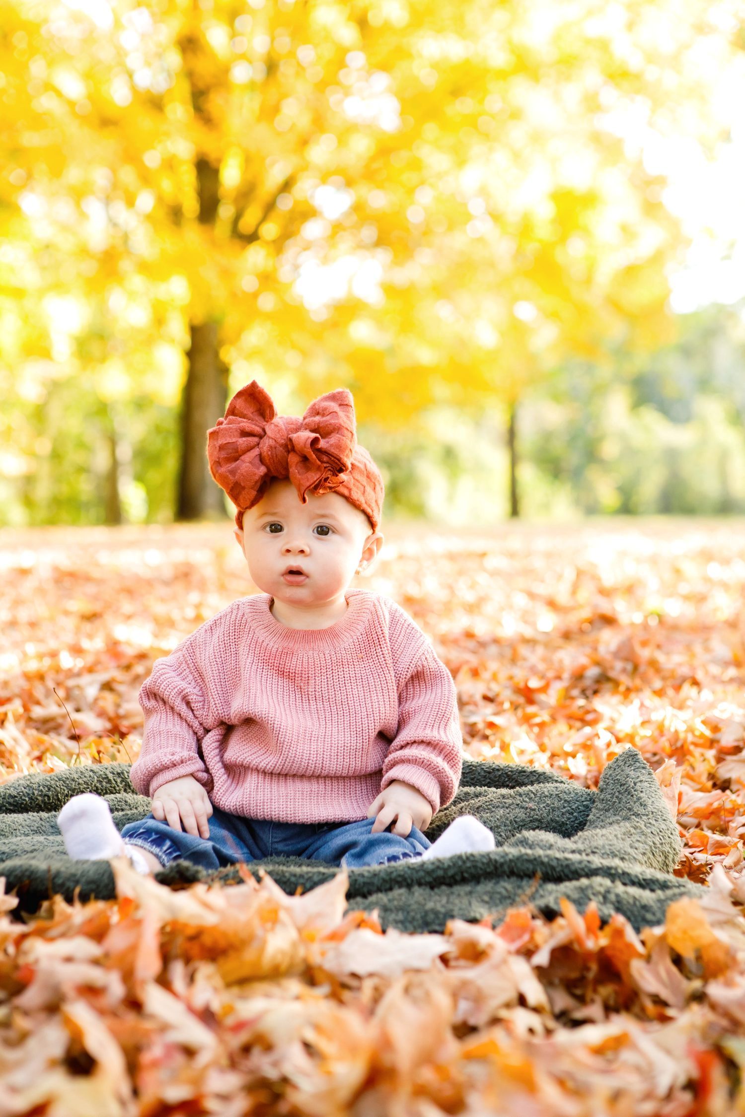 Baby girl in pink sweater and bow headband sitting on blanket in autumn leaves.
