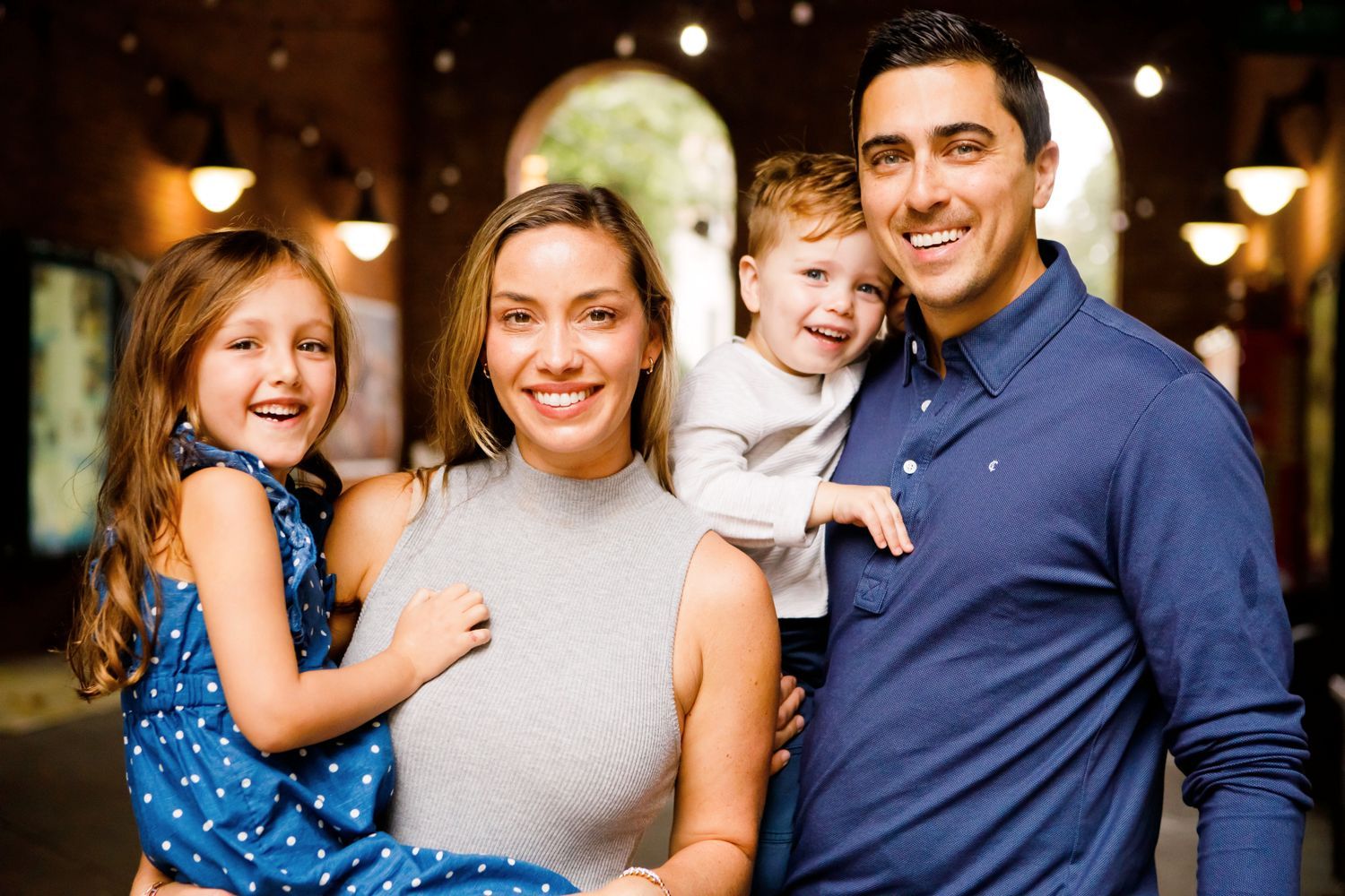 A smiling family of four posing together under warm string lights in an indoor venue.