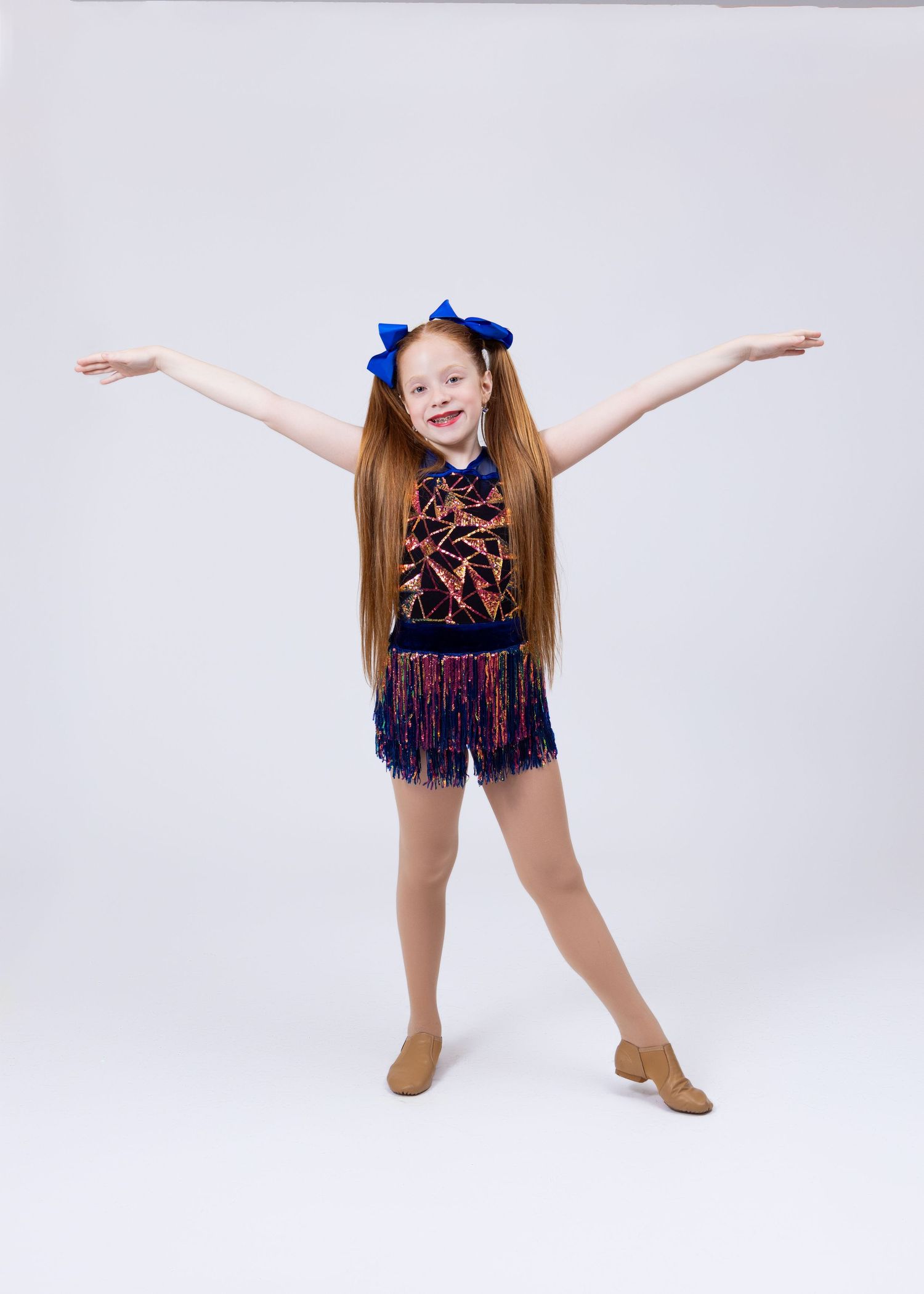 Young red-haired girl in colorful fringe dance costume strikes wide open-arm pose with blue bows.