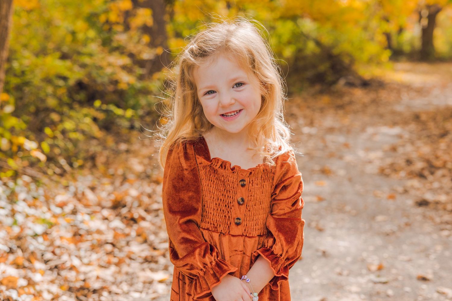 Adorable young girl in rust orange dress smiling on an autumn leaf-covered path outdoors.