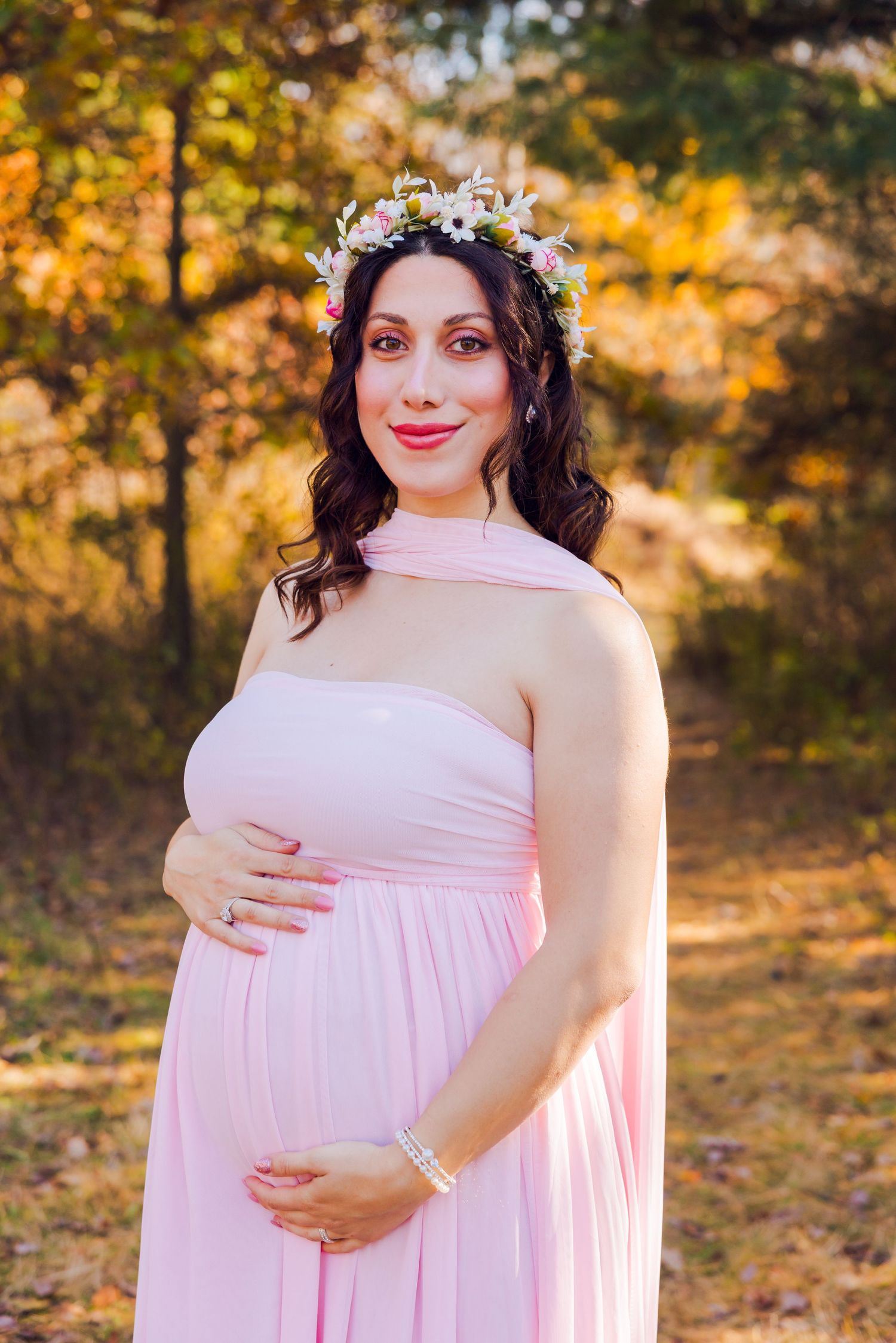 Pregnant woman in pink strapless gown wearing flower crown cradling baby bump in autumn park.