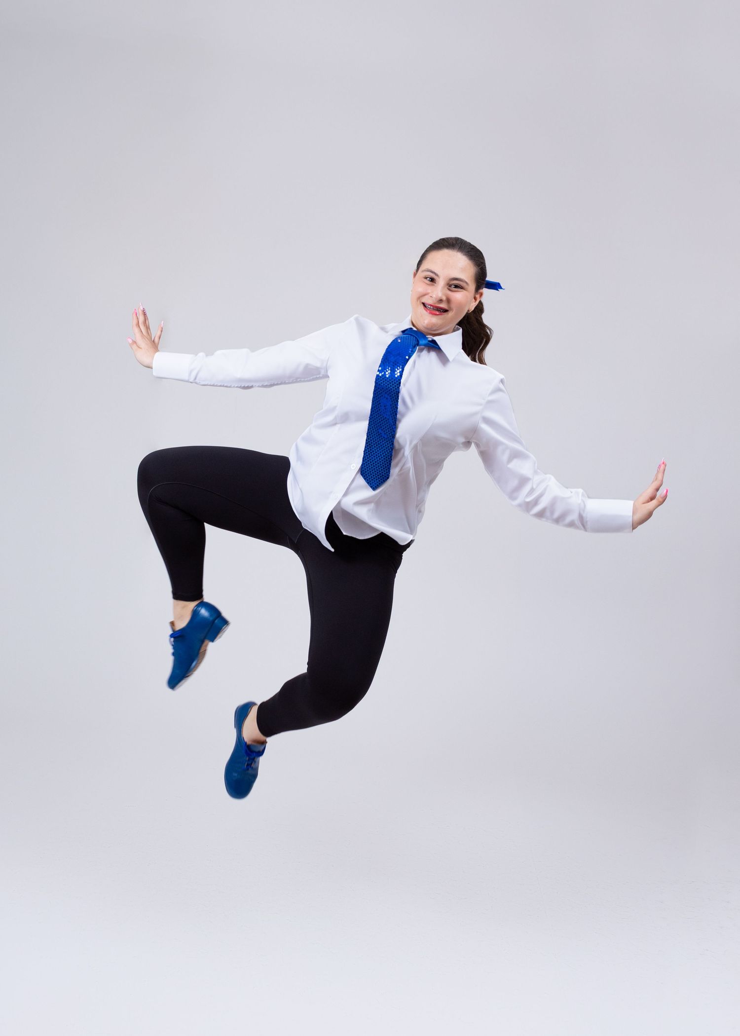 A female dancer in a white shirt and black leggings leaps joyfully in a bright studio setting.