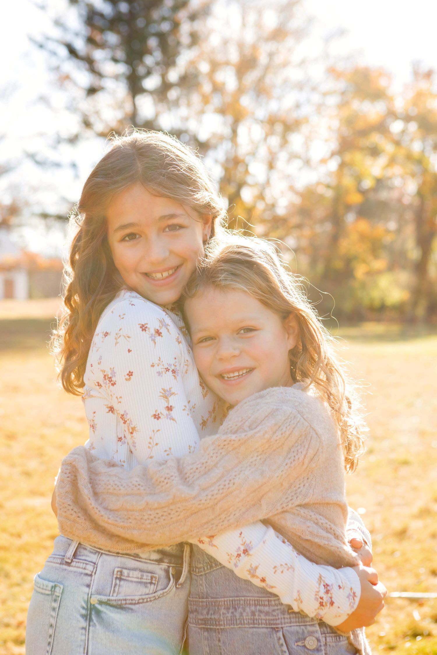 Two young girls hugging each other and smiling in a sunny autumn outdoor setting.