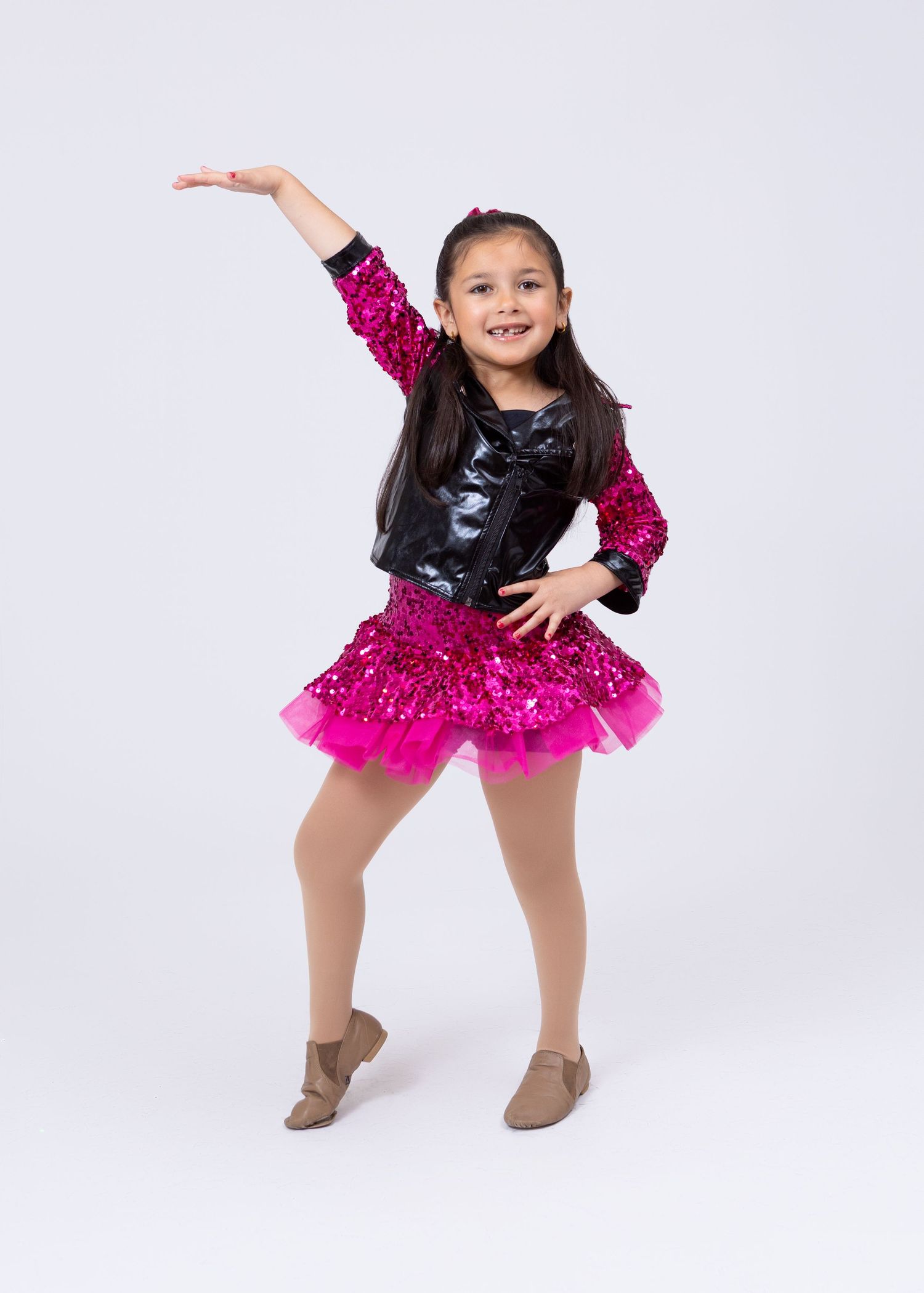 A young girl in a black jacket and pink sequined skirt performs a dance pose on a white background.