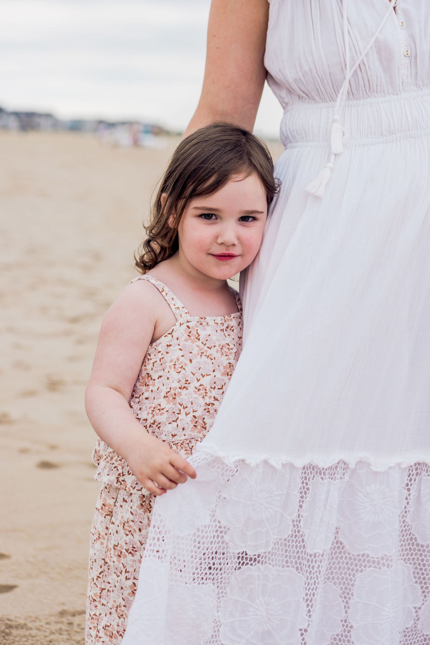Young girl in floral dress hugging her mothers white lace skirt on a sandy beach.