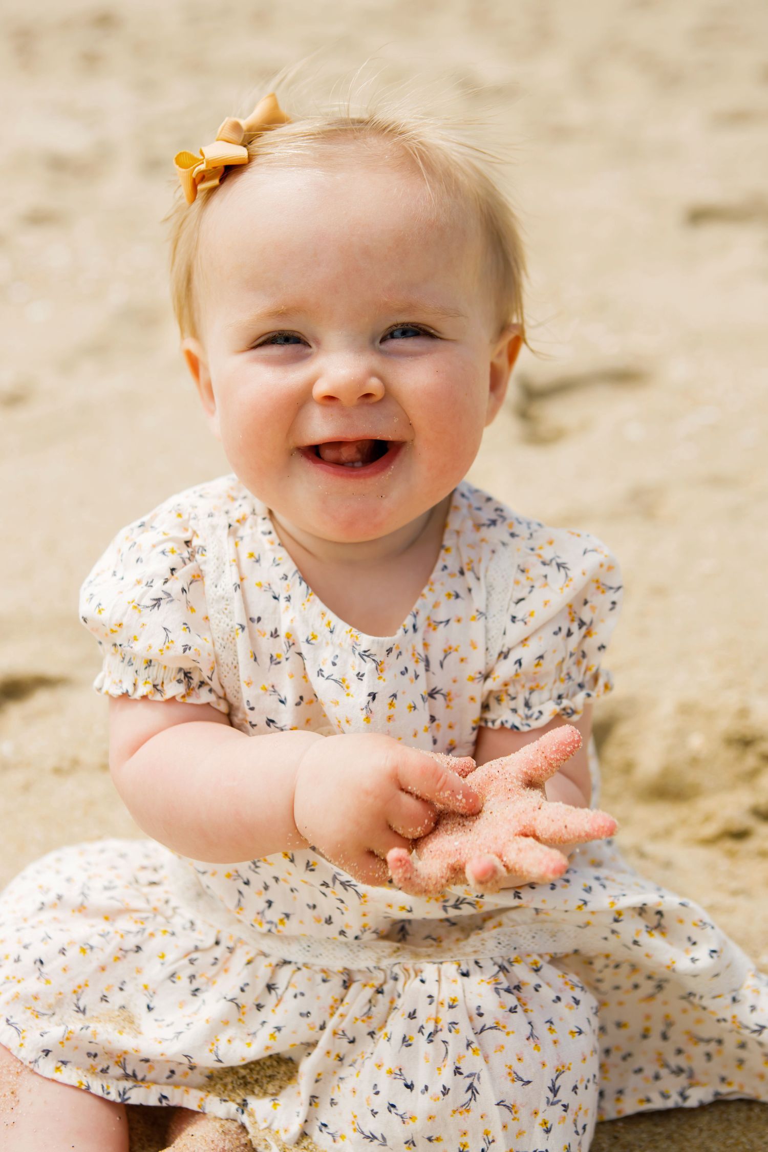An adorable smiling baby girl in a floral dress playing with sand at the beach.