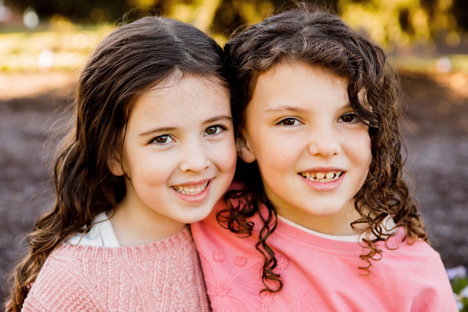 Two young girls with curly hair smiling closely together in a warm outdoor portrait.