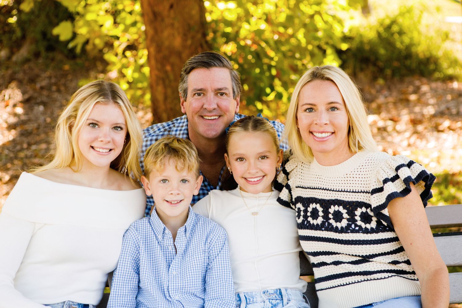 Family of five smiling together outdoors on a bench surrounded by lush green trees.