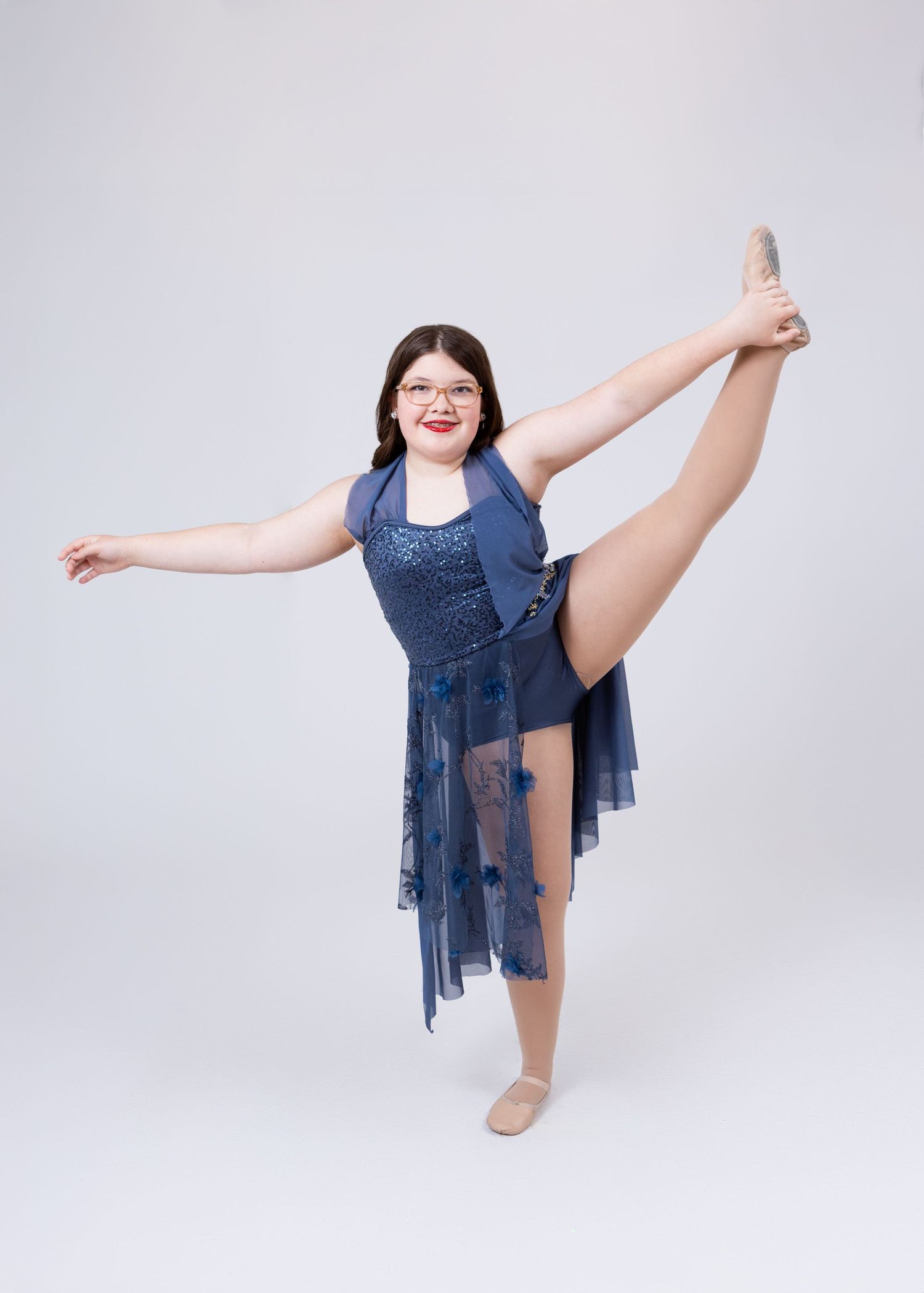 A flexible dancer in a blue sequin lyrical costume performs a standing leg hold pose.