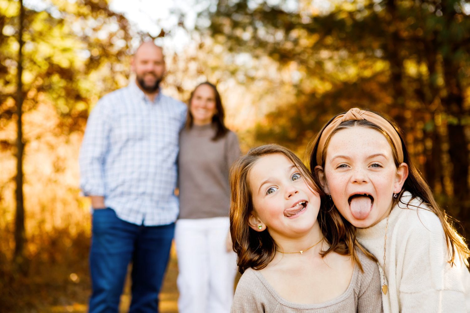 Two girls sticking out tongues playfully with parents smiling in golden fall background.