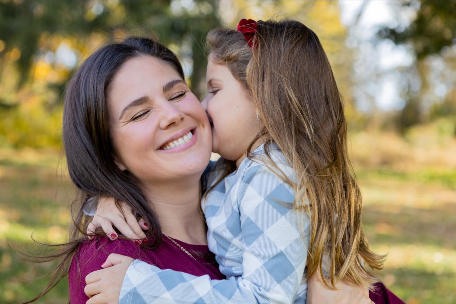 Little girl in blue dress whispers in smiling brunette mothers ear during outdoor fall photo session.