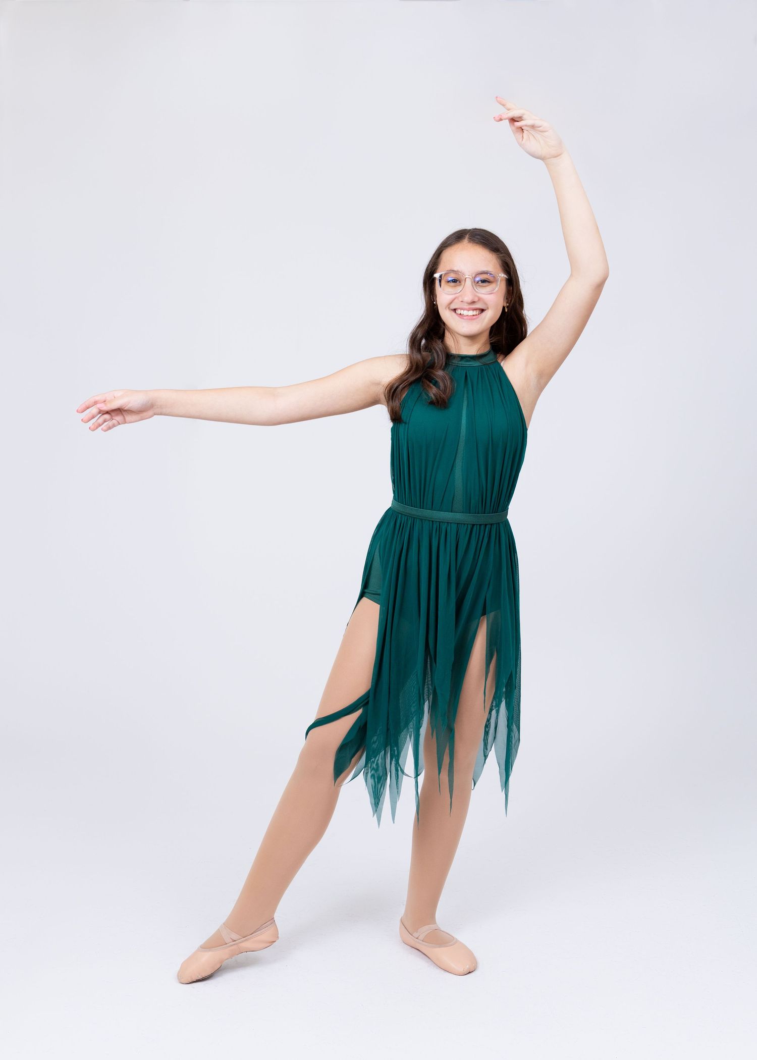 Young girl in emerald green fringe lyrical dance dress performs elegant arabesque pose.