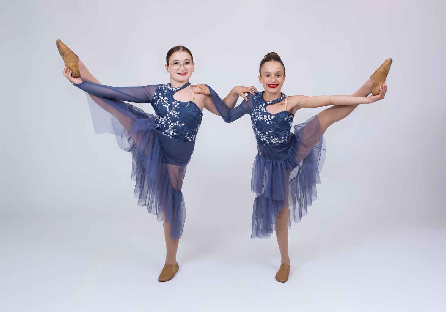 Two young dancers in navy blue costumes perform high leg extensions in a studio portrait.