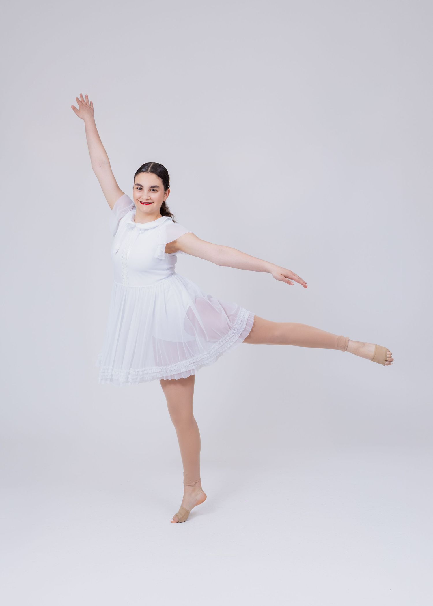 Young dancer in white short-sleeve dress performs arabesque with arms extended gracefully.