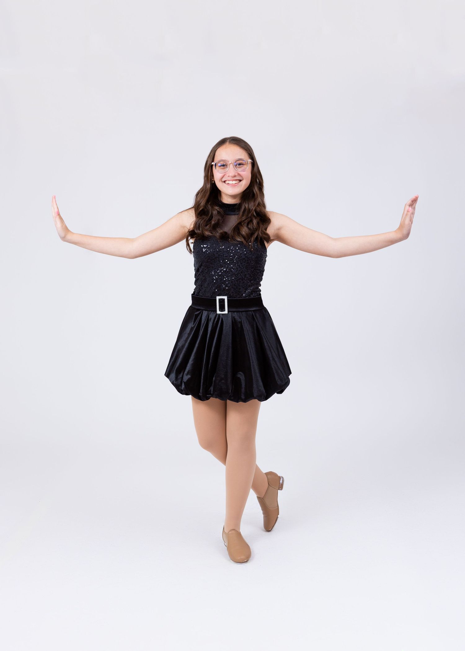 Teen girl in black sequin dance costume with belt performs tap dance pose on one foot.