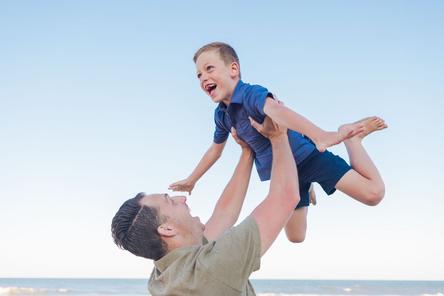 Joyful father lifting laughing young son high in the air on a sunny beach.
