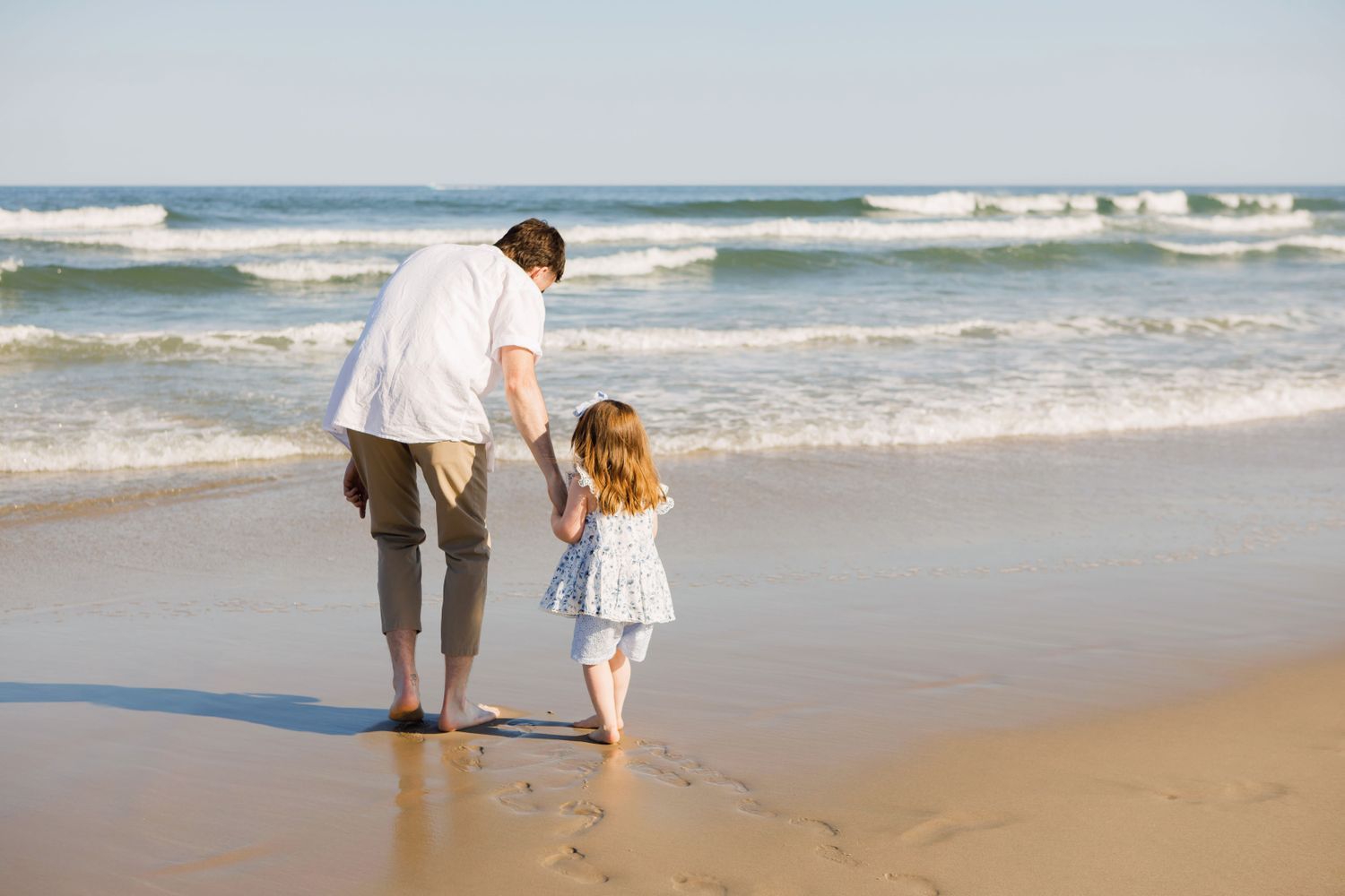 A father and young daughter holding hands while walking along a sunny ocean shoreline.