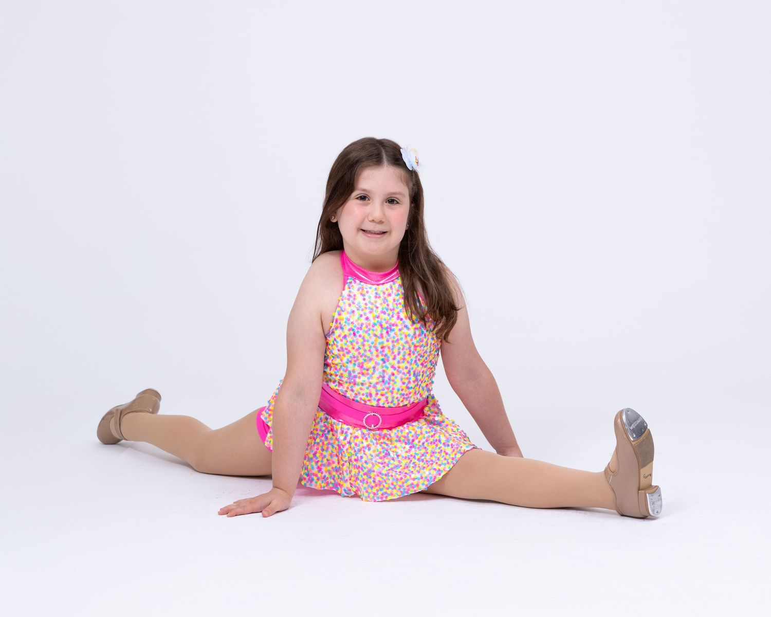 A young girl in a colorful halter dance costume sits in a split pose on a white background.
