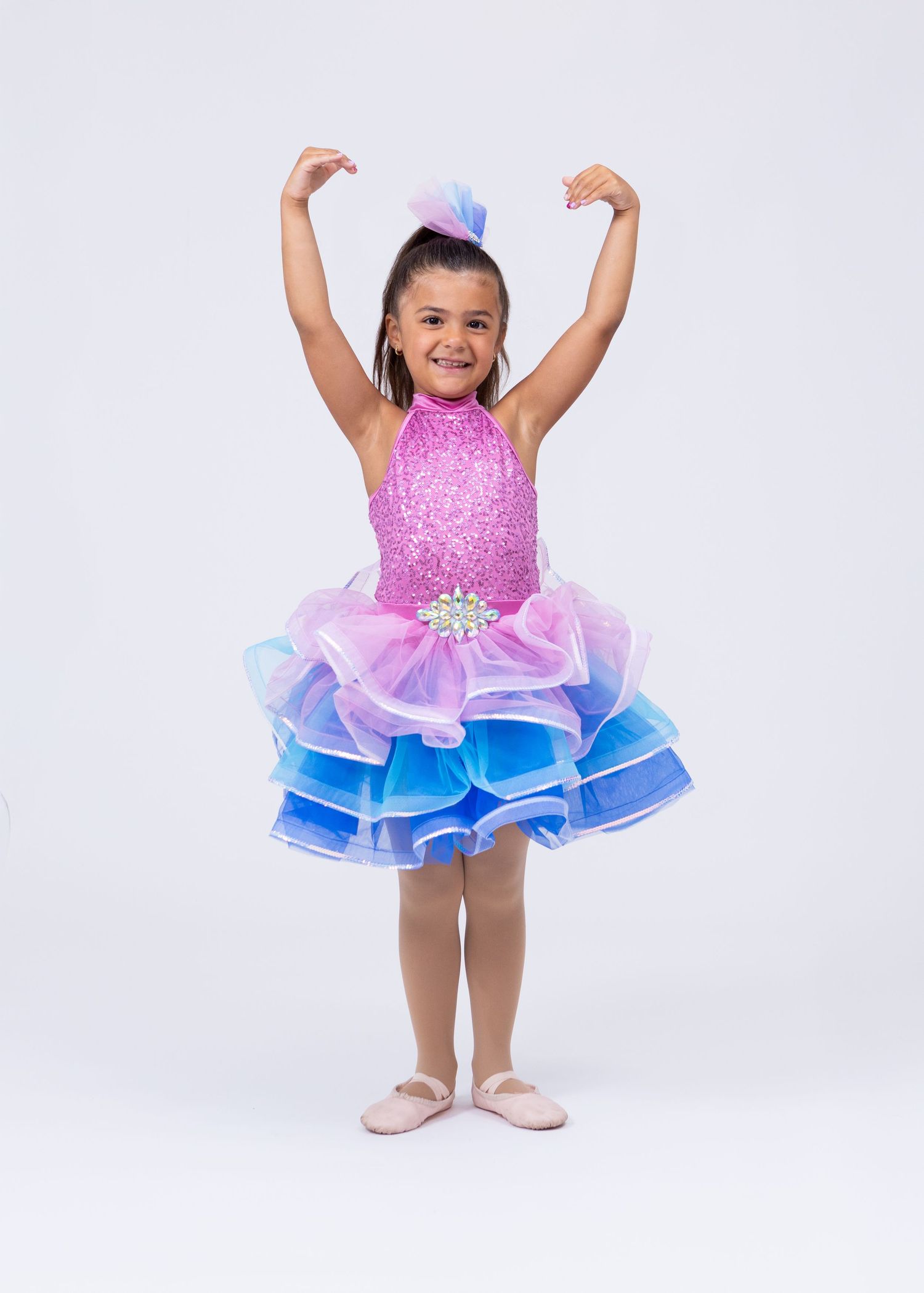 Young girl in pink and blue tiered tutu dance dress with arms raised on white background.